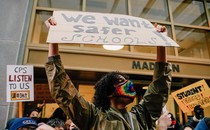 A young man wearing a rainbow mask holds up a sign that says "We want safer schools." In the crowd behind him, other signs read: "CPS Listen to Us" and "Student Voices Matter"