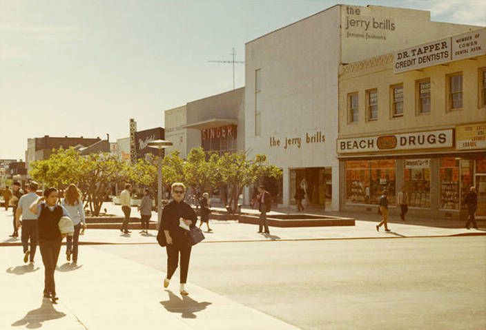 Vintage photo of people shopping on a downtown street. The signs on the front of the businesses read “Beach Drugs,” “The Jerry Brills,” and “Singer.”
