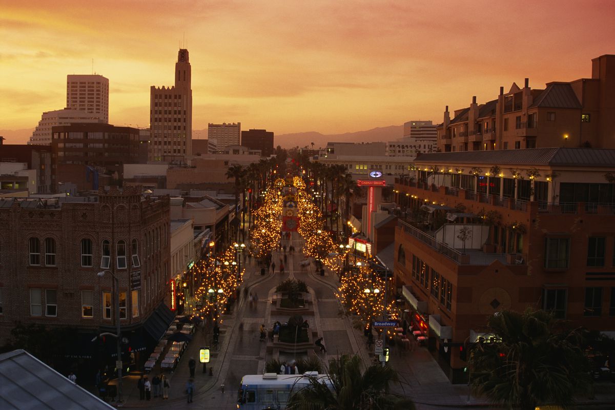 Aerial view of a downtown at sunset, with tall and mid-size buildings lining a wide boulevard open only to pedestrians. Hills are present in the background.