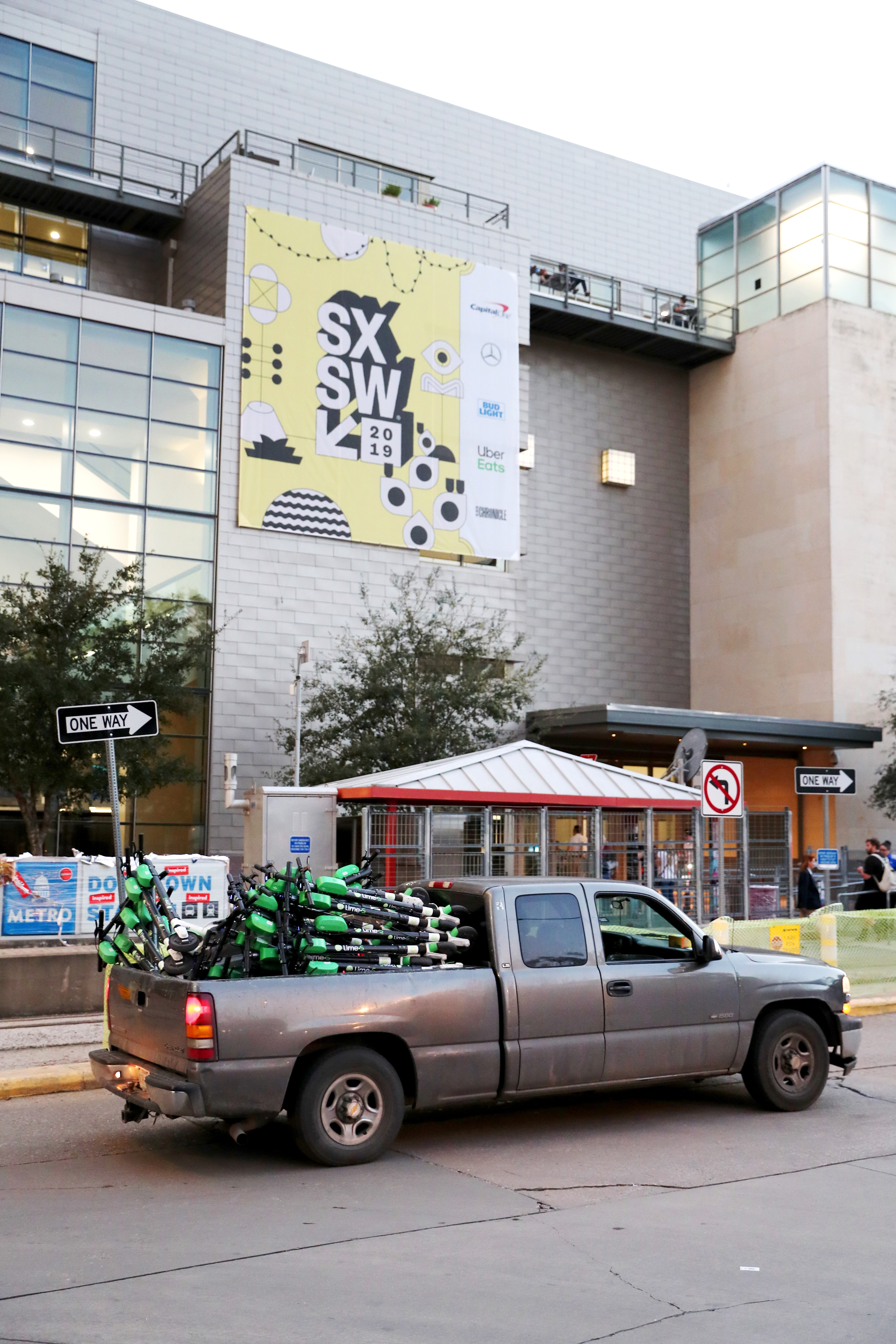 A truck full of e-scooters is parked on an asphalt street  in front of a small, covered commuter rail station. A five-story building with sign that reads SXSW is in the background. One-way street signs are on the sidewalk. 