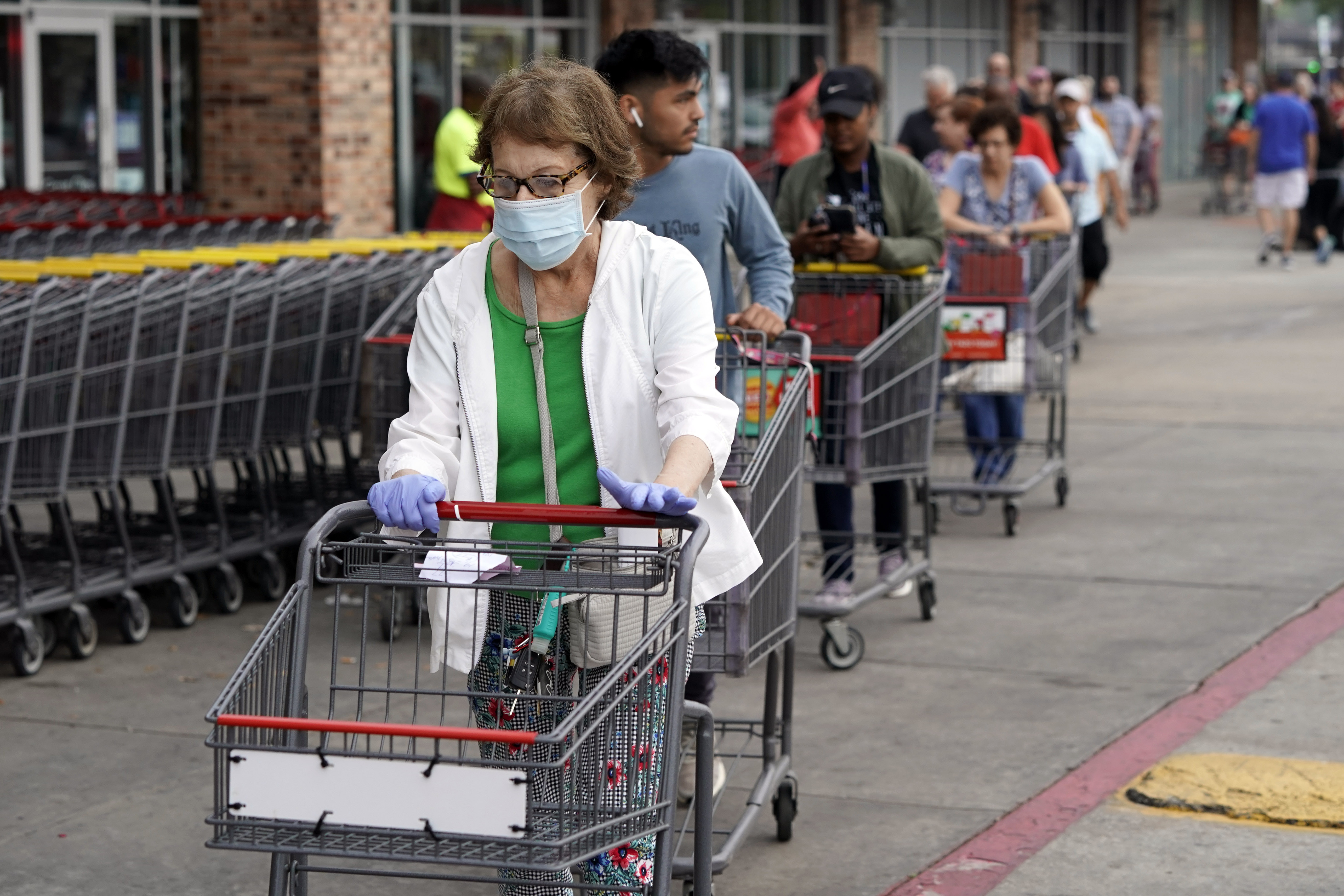 A white woman with brown and gray hair wears a mask and gloves as she enters an H-E-B grocery after waiting in line with more than 150 people. She’s pushing a grocery basket.