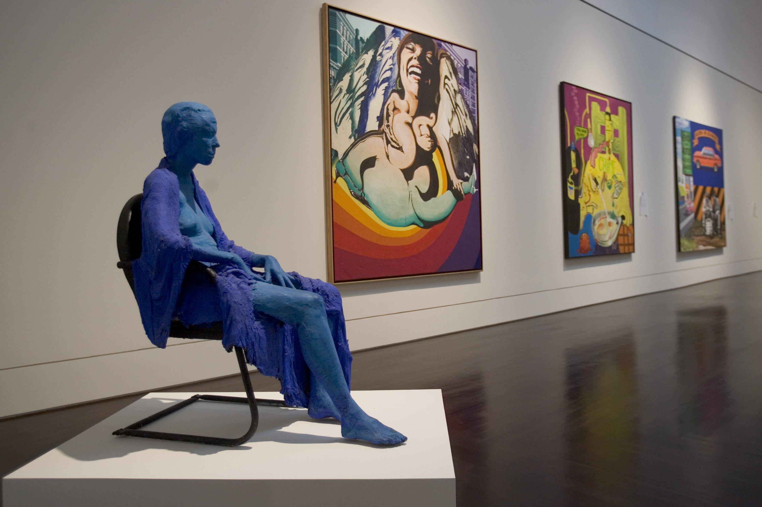 A sculpture of a blue woman sitting in a chair in an art gallery with colorful pop-art paintings hung on the white wall.