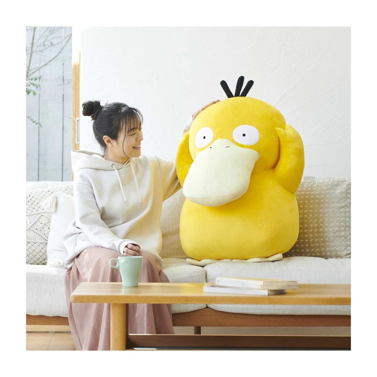 A woman sitting on a couch next to a life-sized Psyduck plush toy. She’s rubbing it’s big yellow head as it gazes into the distance.