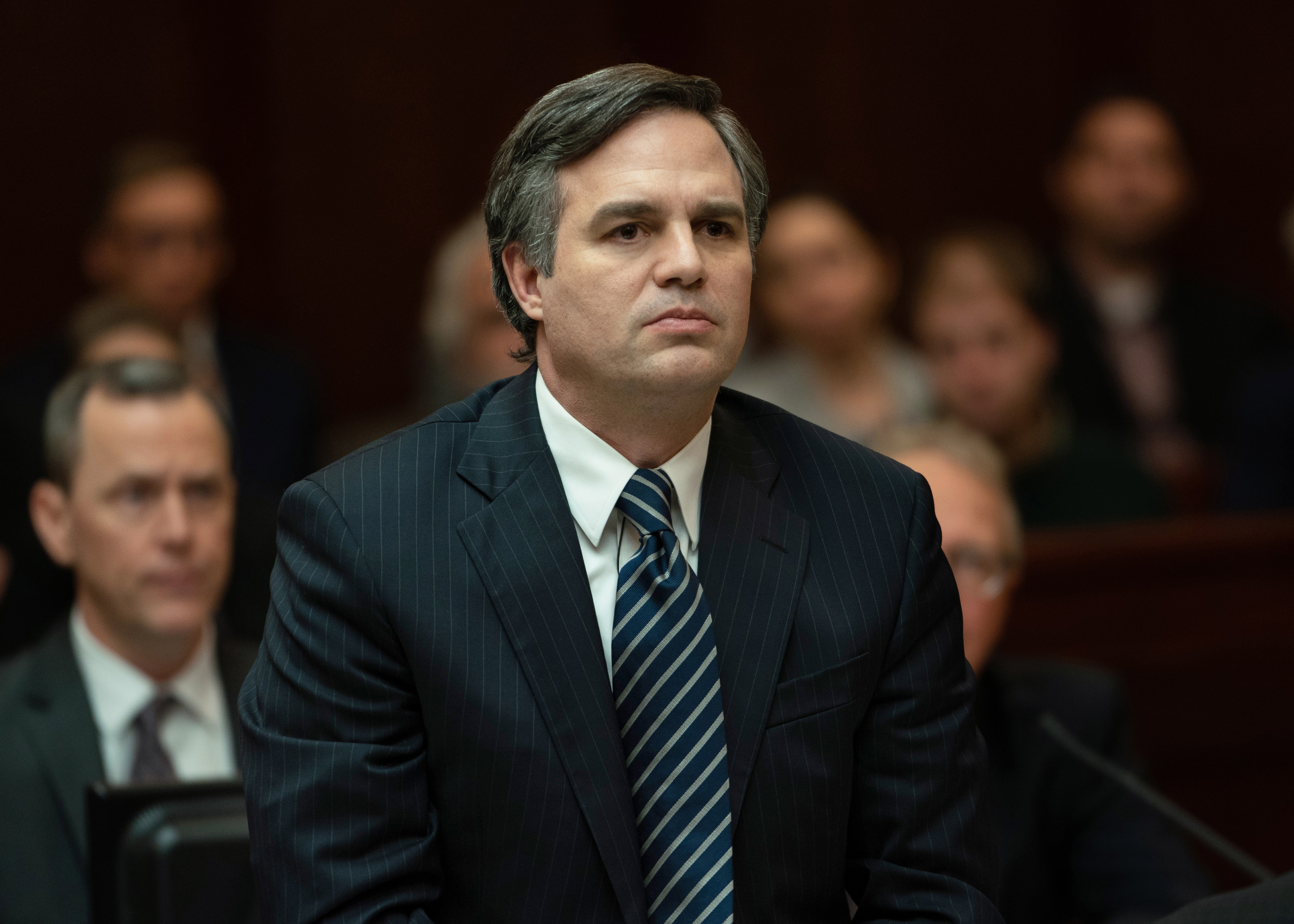 A medium close-up shot of a man wearing a dark blue suit with a striped necktie sitting at the witness stand of a courtroom in Dark Waters.