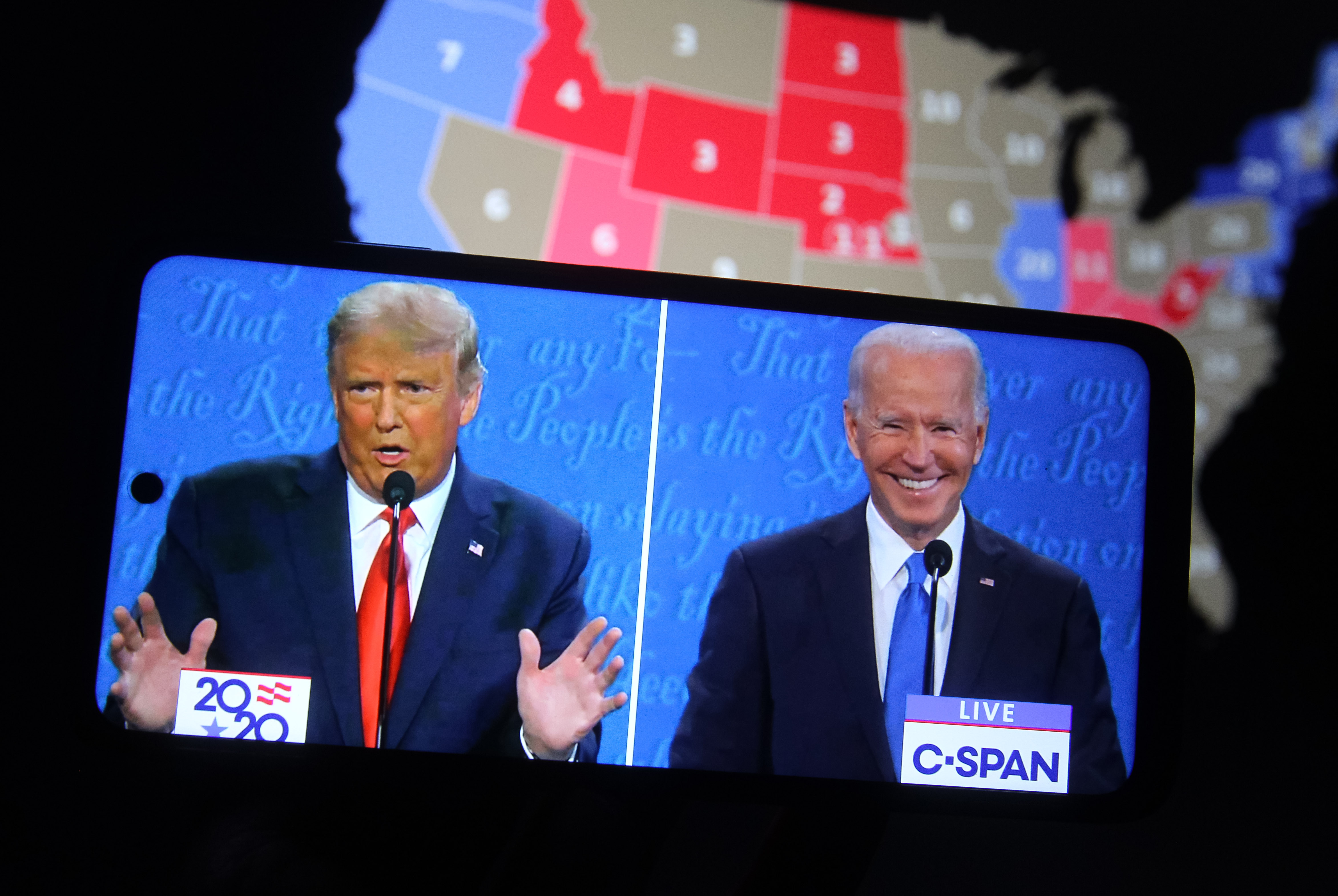 Donald Trump and Biden pictured side-by-side on a phone screen, speaking into podium microphones. Behind the phone is a map of the United States.