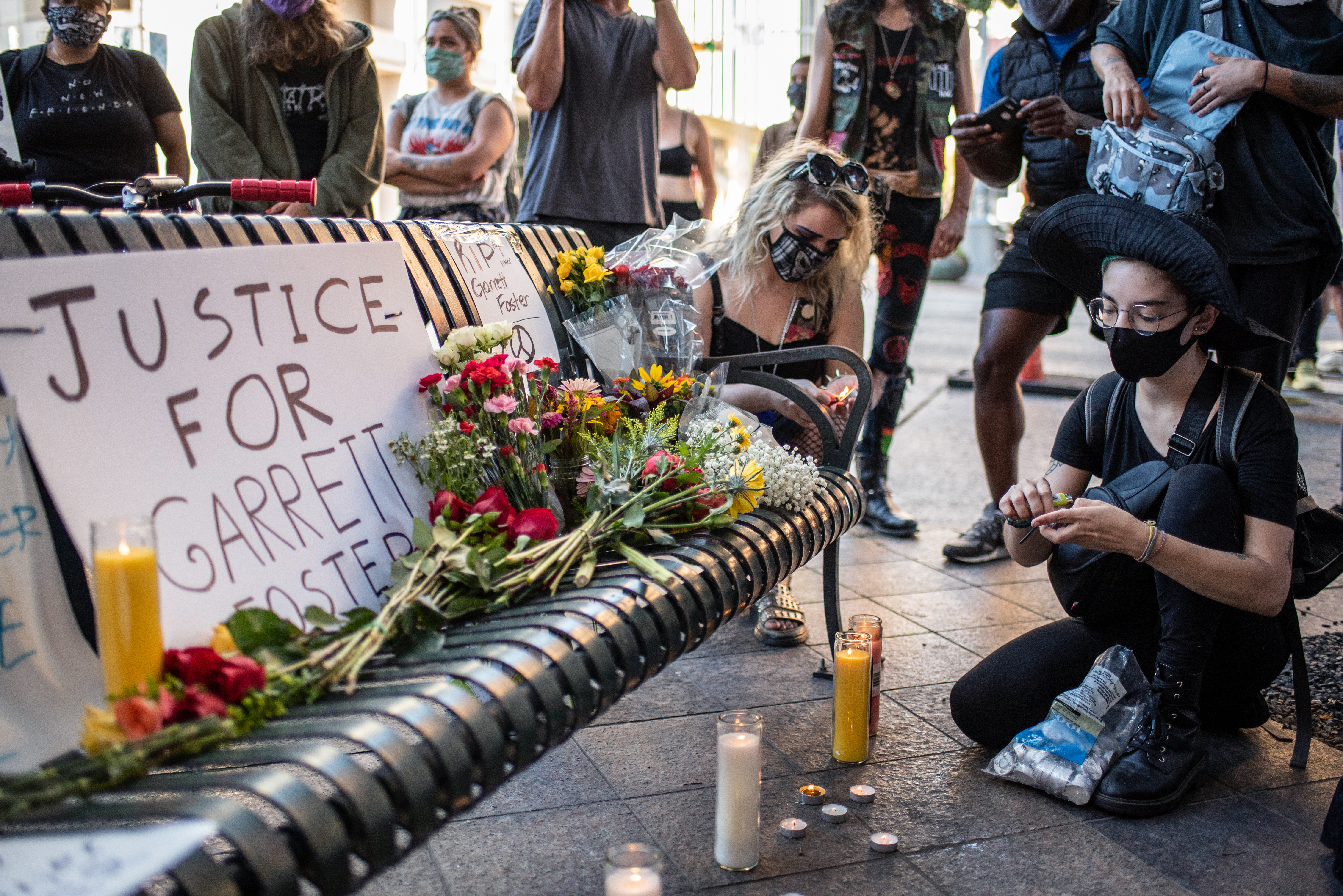 People light candles and kneel around a metal bunch with a sign reading “Justice for Garrett Foster” and several flower bouquets on it.
