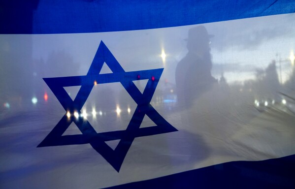 A demonstrator crossing the street is silhouetted behind a flag of Israel during a rally in support of Israel Monday, Oct. 9, 2023, in Bellevue, Wash. (AP Photo/Lindsey Wasson, File)