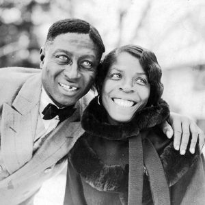 African-American man in suit who has a gap in his teeth with his arm around a smiling woman wearing a fur-trimmed coat