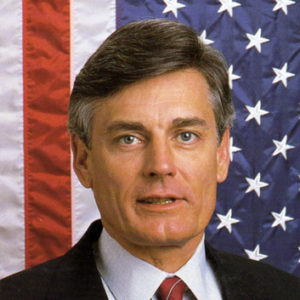 white man in suit and tie with American flag backdrop