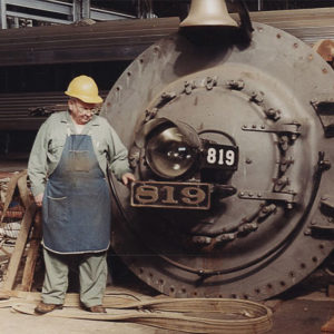 White man in hardhat and work clothes standing before locomotive components