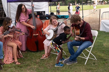 Three women and one man sit in folding chairs in a half circle on grass. They are playing instruments like the fiddle, the upright bass, the banjo, and the acoustic guitar. A child puts his ear up to the body of the guitar as the man plays, and puts his fingers out to feel the vibration of the strings.