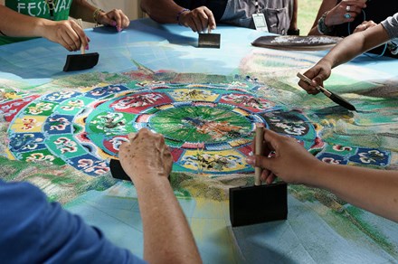 An intricate Tibetan Buddhist sand mandala sits on a blue tabletop. It has been segmented into slices, and five people reach their arms over top of it with brushes to disperse the sand into the air.