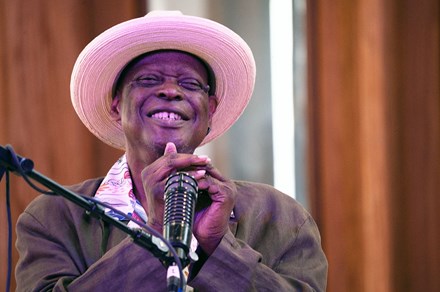 An elder Black man smiles on stage, cupping a harmonica near a microphone.