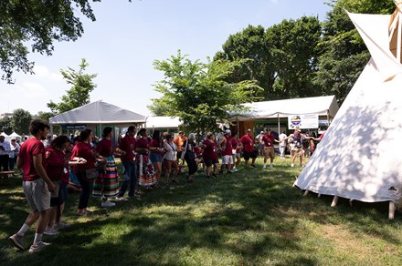 Several people, many in matching red shirts, form a circle around a white tipi outdoors.
