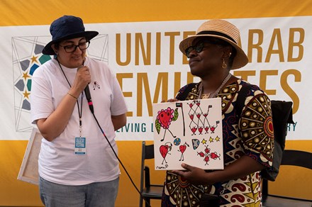 A woman speaks into a microphone as another woman next to her smiles, eyes closed, holding a piece of artwork depicting red hearts with eyes and smiles, some holding UAE flags.