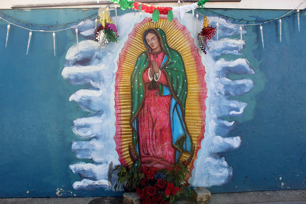 Painting of the virgin on an outdoor wall, with red flowers placed at her feet, and string lights draped above.