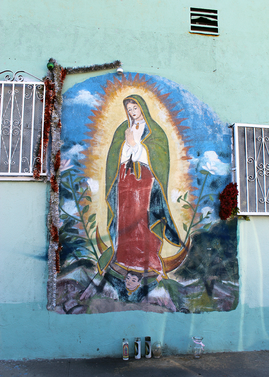Painting on the side of a pale blue building, between barred windows. It features the virgin in red skirt, green shawl, and white flowers around her. Set on the ground below her and candle holders.