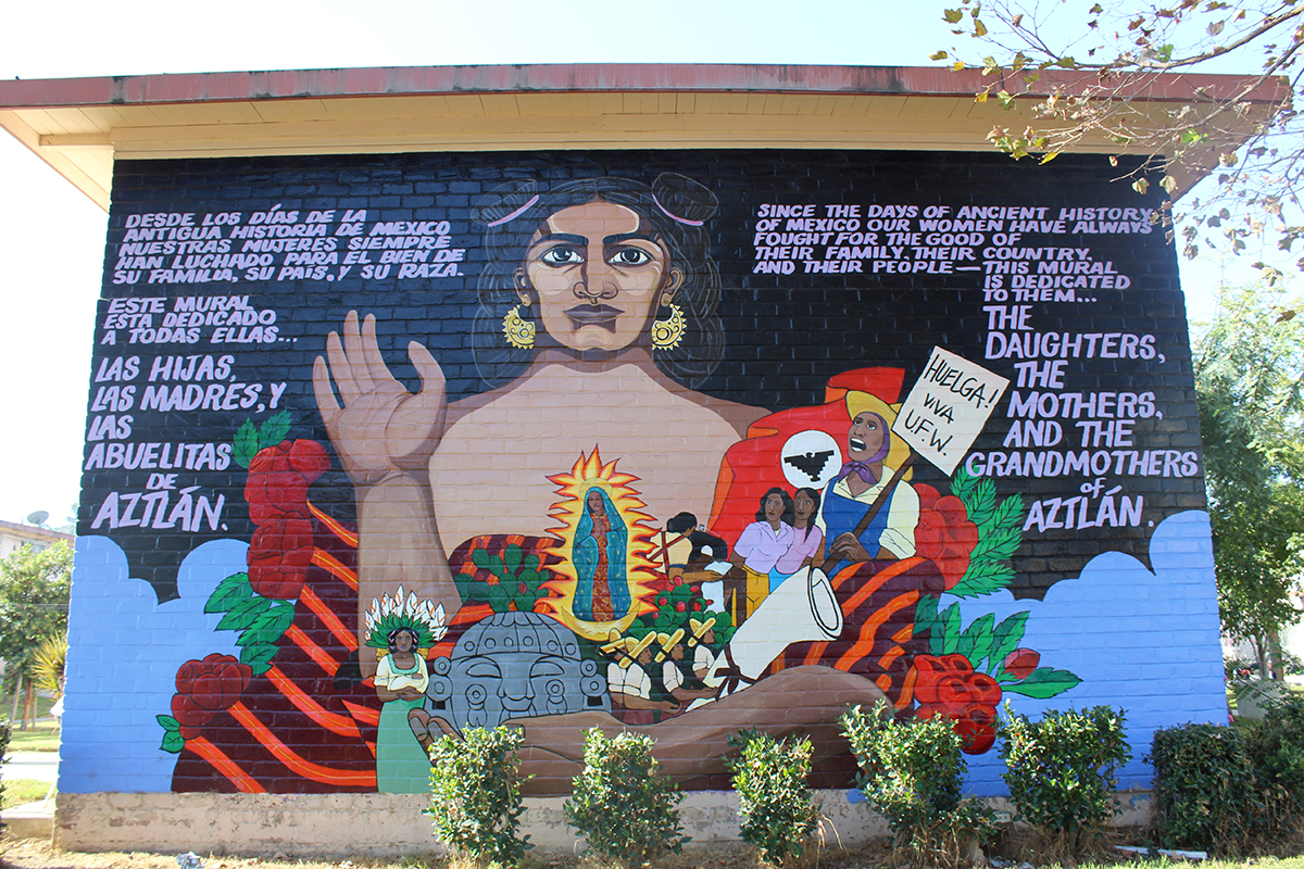 Mural on the side of a brick building, featuring a looming brown-skinned figure with gold earrings and one open hand raised. In their other arm, the person holds smaller figures, including Guadalupe, farmworkers with a Huelga! VIVA UFW sign raised, workers holding a giant diploma, red roses, and gray stone artwork of a face. Text on the left in Spanish, and the English translation on the right: Since the days of ancient history of Mexico our women have always fought for the good of their family, their country, and their people. This mural is dedicated to them... the daughters, the mothers, and the grandmothers of Aztlan.