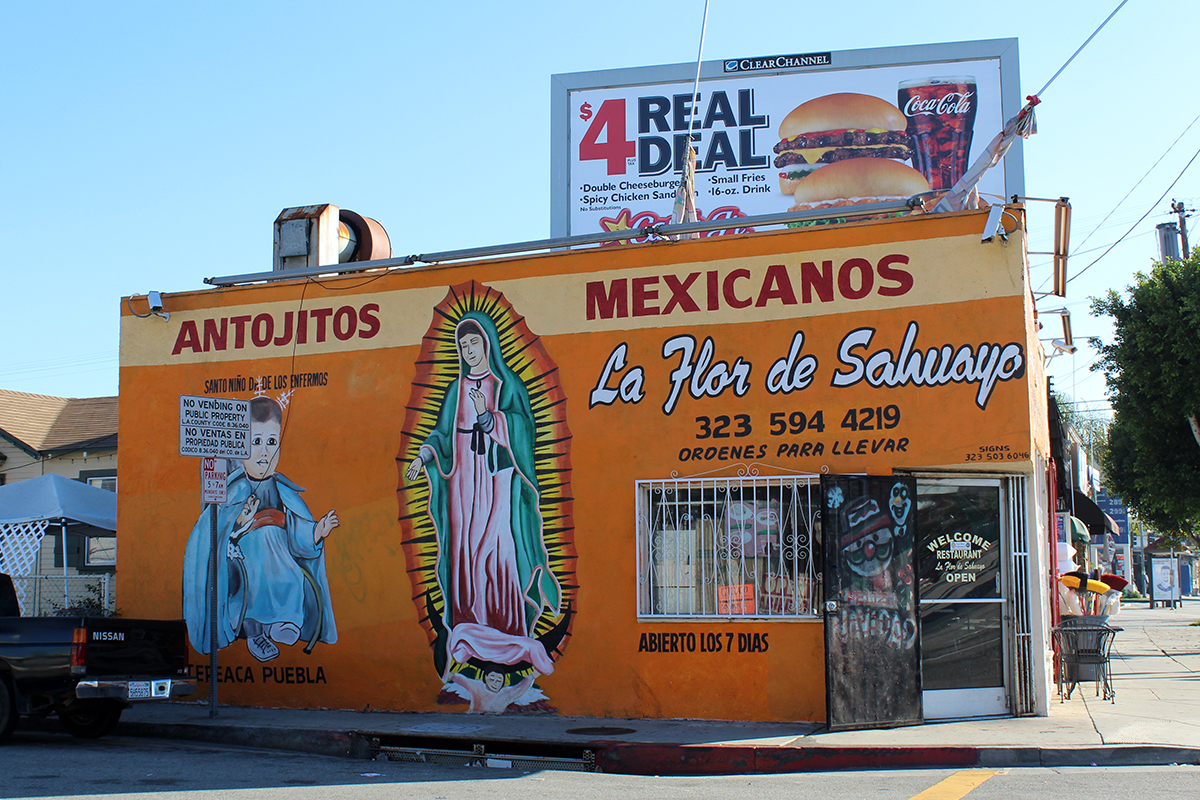 Mural on the side of an orange building, feature a virgin figure, a baby saint figure, and words in Spanish: Antojitos Mexicanos. La Flor de Sahuayo. Above and behind the building, a Carl's Jr. billboard with a burger and a Coke.