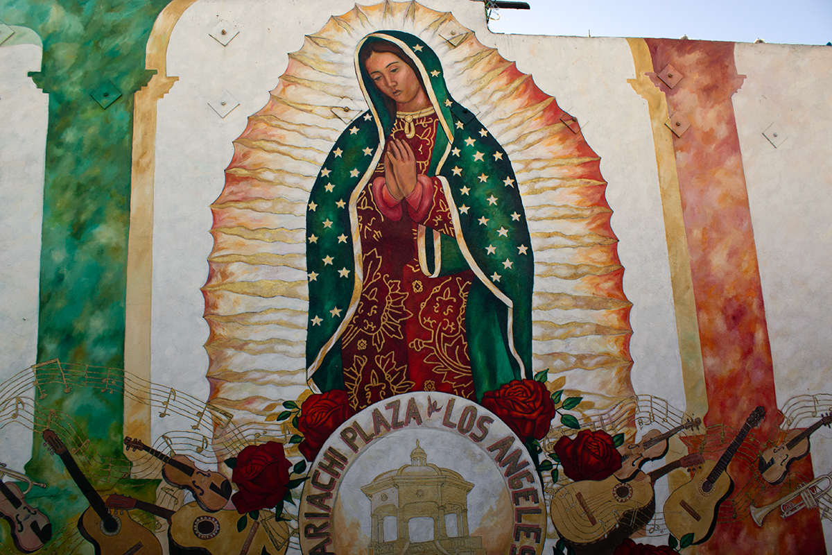 Painting of Guadalupe in red, gold, and green, with red roses, guitars, violins, and trumpets painted at her feet. A circular emblem at the bottom reads Mariachi Plaza de Los Angeles.