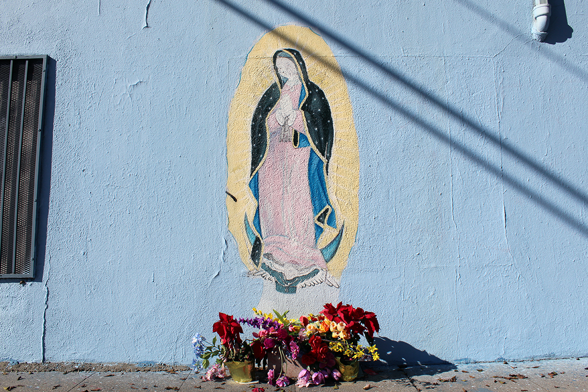 Painting of the virgin on an exterior pale blur wall, with multicolored flowers set on the ground below her.