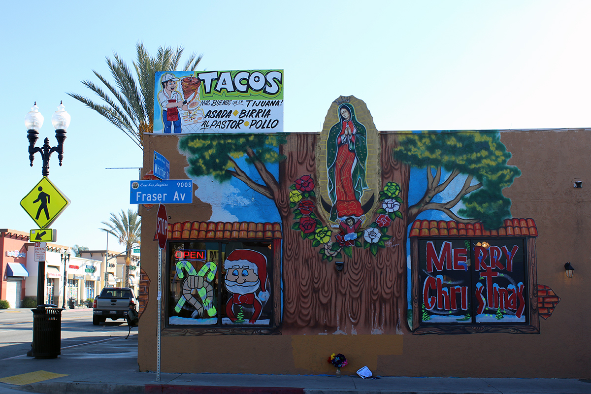 A corner store with a sign for Tacos on top. A painting on the side includes the virgin set on top of a wide tree trunk, with Santa Claus and the words Merry Christmas painted on the windows.