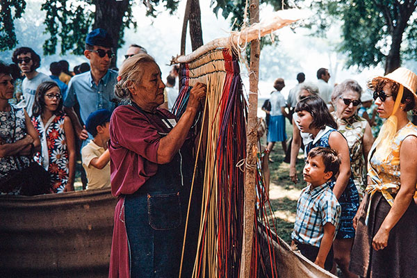 A person leaves a mat hanging from a standing wooden frame while a small crowd of children and adults watch. Old color photo.