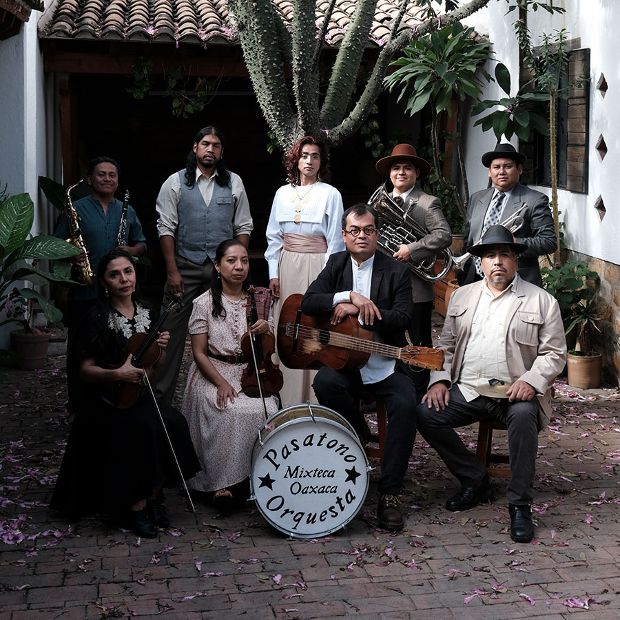 Eight adults pose in front of an adobe brick wall holding musical instruments: violines, clarinet, double bass, acoustic guitar, horn, small guitar, and drum.