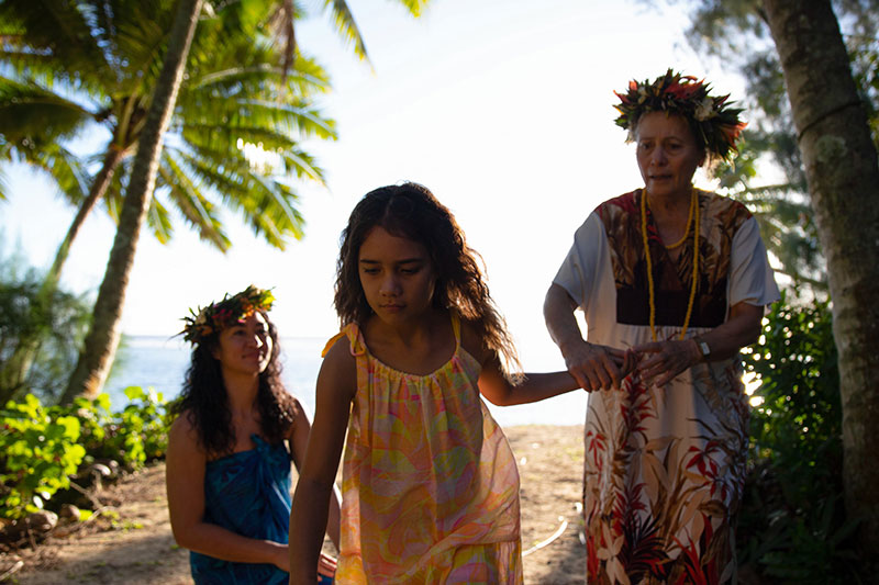 A young girl walks away from two women, the older of whom is holding her hand, with palm trees, beach, and ocean in the background. All three are in floral dresses.