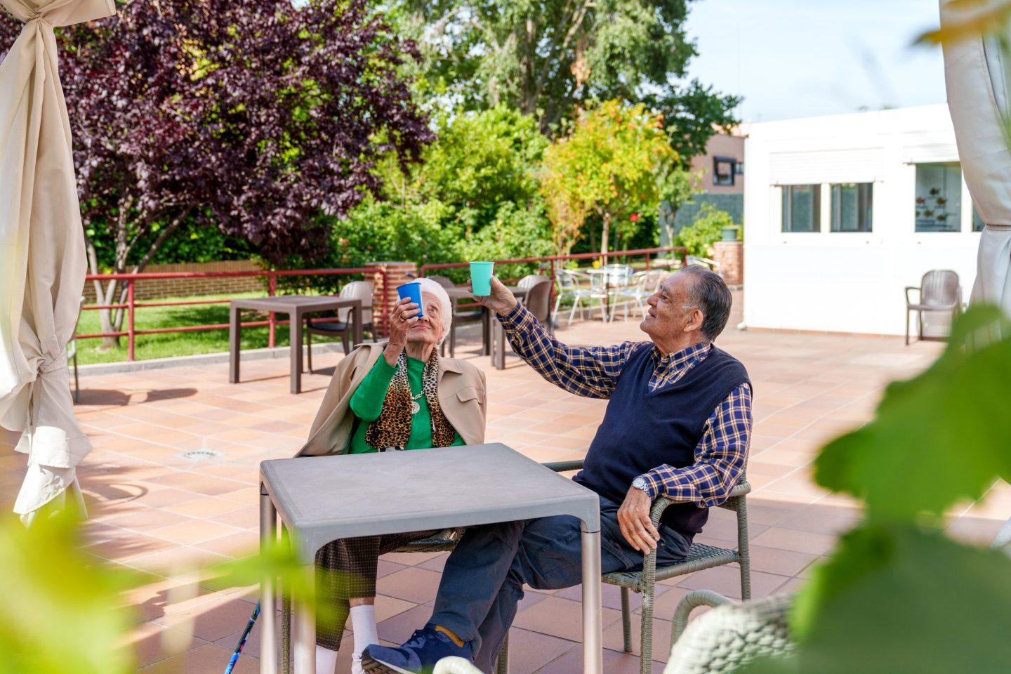 Joyful elderly man and woman sitting in a well-manicured garden at a senior care facility, happily toasting with vibrant glasses on a sunny day.