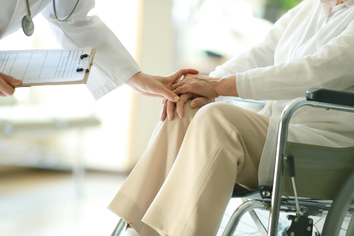 Older person sits in wheelchair as doctor places hand on theirs.