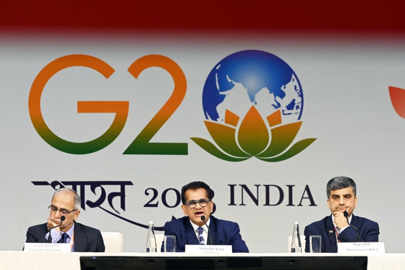 Three men wearing suits and ties sit at a table in front of a wall displaying the logo of the New Delhi G-20 summit, which stylizes the zero in G-20 as the globe. The man sitting in the middle has his mouth open as he speaks into a microphone.