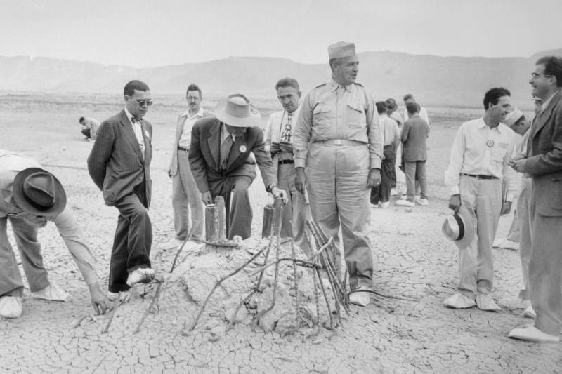 A historic black-and-white image from the aftermath of the first atomic bomb testing. Scientist J. Robert Oppenheimer stoops to examine the torn and warped metal remaining from the base of a tower from which the bomb was tests. Other scientists mill about the desert landscape around Oppenheimer, and low mountains loom in the distance.