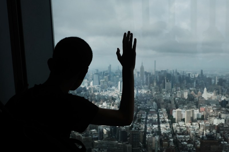 A teenager is shown from behind as a silhouetted figure as he looks out at Manhattan from a window at the top of One World Trade Center. His hand is raised at a right angle with his fingers pressed against the glass. The New York City skyline is visible through the glass beneath a cloudy sky.