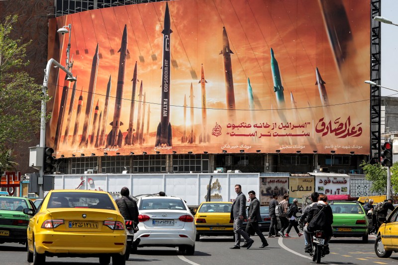 Motorists and pedestrians travel on a paved road beneath a massive billboard in Tehran. The billboard depicts a fleet of missiles standing upright against a background of orange flames, along with text in Persian and Arabic.
