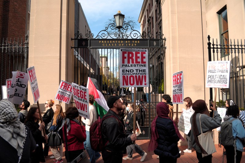 People with Free Palestine posters walk past a campus gate that reads "PRESENTED BY THE CLASS OF 1929"