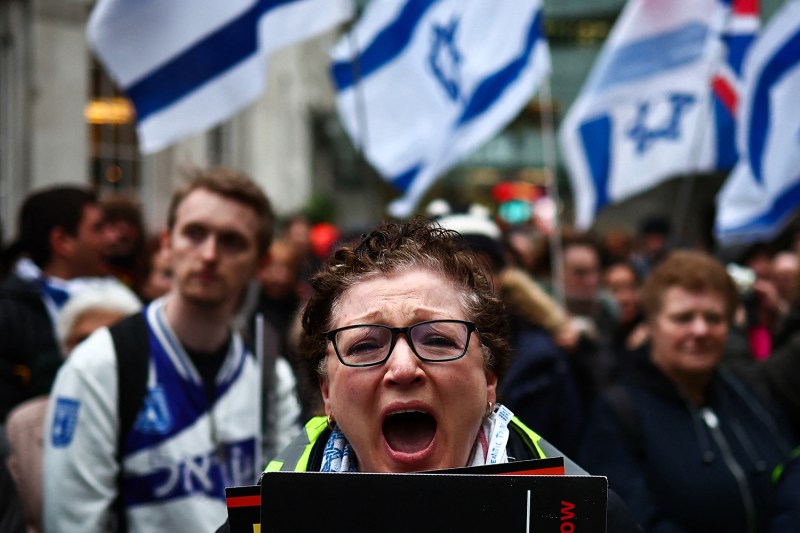 A protester wearing glasses and holding a sign screams into the camera. Behind is a crowd of more protesters holding signs and waving Israeli flags.