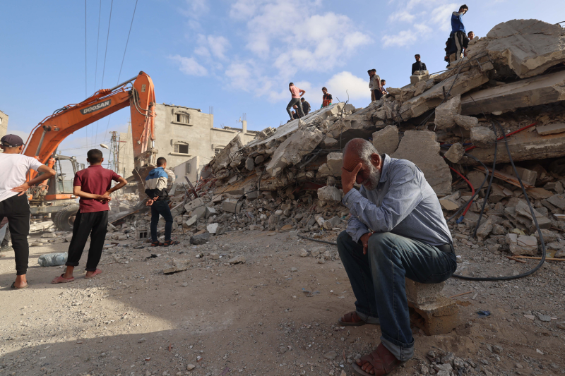 A Palestinian man sits on a piece of rubble beneath a blue sky with his head cradled in his hand as he waits for news about his missing daughter. In the background, rescue workers climb a stack of debris from a collapsed building as they search for survivors.
