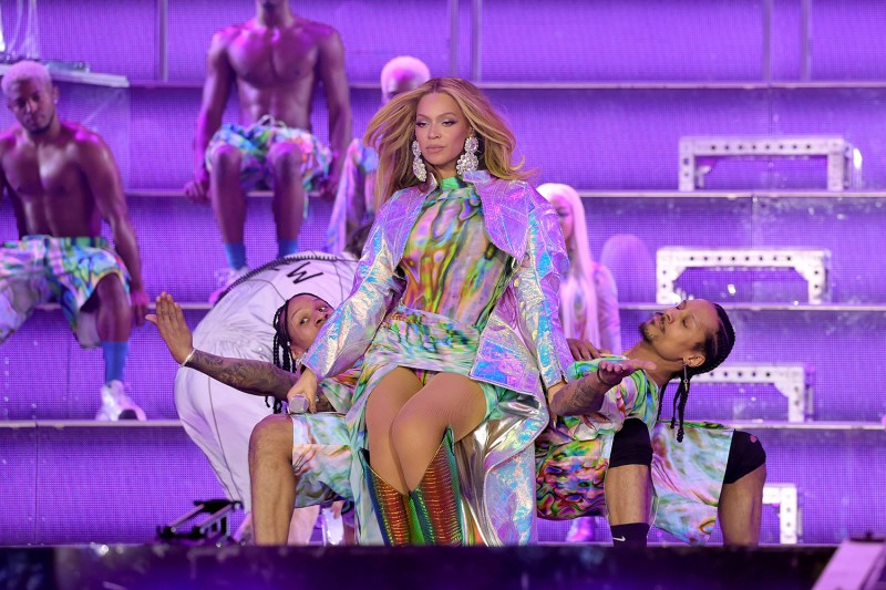 American singer Beyoncé performs onstage at an arena in Sweden, surrounded by background dancers. Beyoncé wears large earrings and a short dress and elaborate jacket made out of the same holographic, silvery fabric. Two backup dancers kneel in bridge poses, and Beyoncé sits on top of them with her microphone as she looks out at the audience with a small smile.