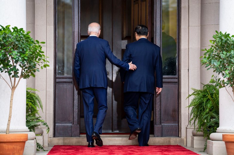 U.S. President Joe Biden and China's President Xi Jinping, both wearing dark suits, are seen from behind as they walk through a large wooden doorway. Biden reaches out to pat a hand on Xi's back. Small trees flank the entrance.