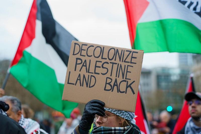 A person with a black glove holds up a sign that reads "DECOLONIZE PALESTINE LAND BACK."