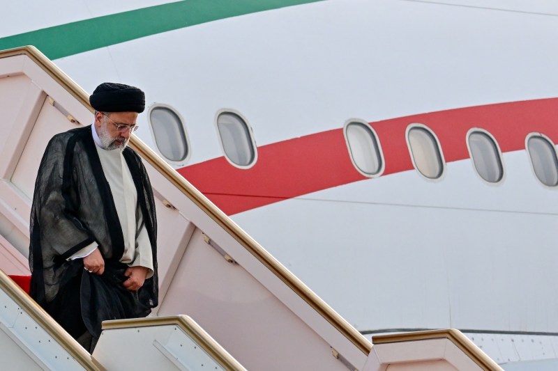 Iranian President Ebrahim Raisi walks down a flight of steps as he exits his plane upon arriving in Sri Lanka. Raisi is a man in his 60s with a white beard and glasses. He wears a black turban and a sheer black robe over an off-white shirt. The side of the plane is white, with stripes in the red and green of the Iranian flag.