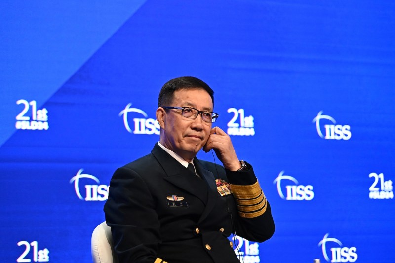 Chinese Defense Minister Dong Jun sits onstage in front of a bright blue wall as he attends the Shangri-La Dialogue in Singapore. He wears a black military dress uniform and glasses, and one hand is raised to his face to adjust his microphone wire.