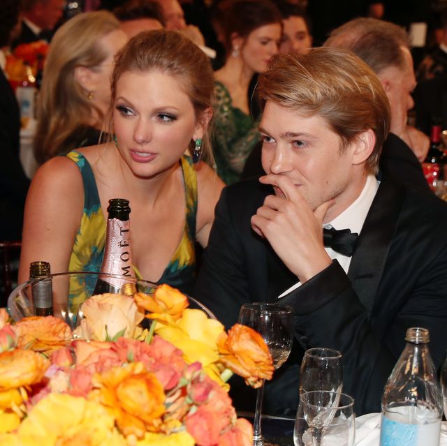 taylor swift and joe alwyn sit at a table with yellow and orange flowers and several drinks on it, she wears a sleeveless blue and orange dress, he wears a black tuxedo and bowtie, they look to the left