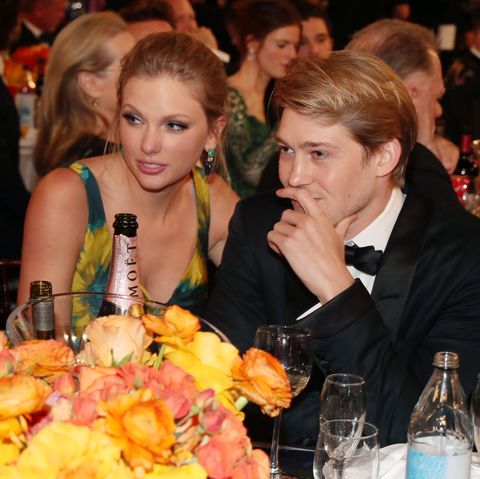 taylor swift and joe alwyn sit at a table with yellow and orange flowers and several drinks on it, she wears a sleeveless blue and orange dress, he wears a black tuxedo and bowtie, they look to the left