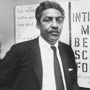 bayard rustin looks at the camera while standing in front of antisegregation signs, he wears a suit jacket, collared shirt and tie and holds a cigarette in one hand