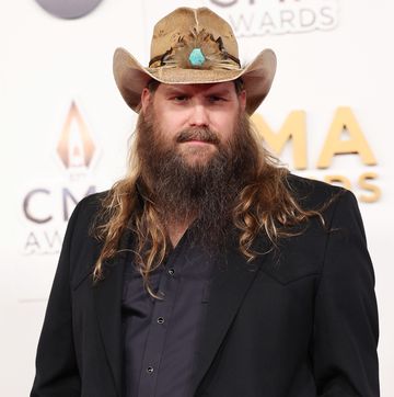 chris stapleton looks at the camera, he wears a black suit jacket, black shirt and light brown decorated cowboy hat