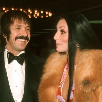 sonny and cher stand next to each other in formal attire, he smiles and she looks left with a small smile