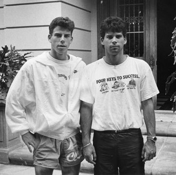erik and lyle menendez stand outside a home and stare at the camera, the front door is open, erik wears a jacket over a tshirt with shorts, lyle wears a tshirt with pants