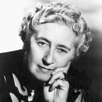 agatha christie looks at the camera as she leans her head against on hand, she wears a dark top and rings on her fingers